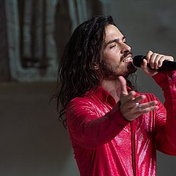 ein Mann mit langen Haaren und ein rotes Hemd singen in ein Mikrofon